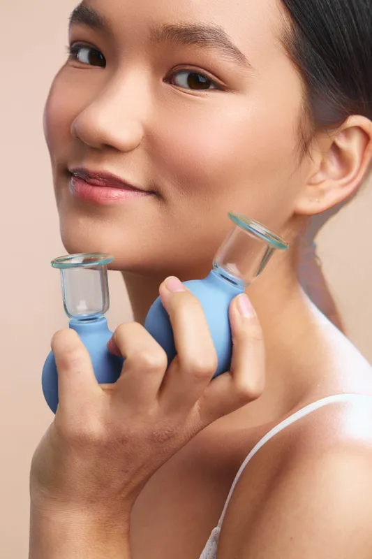 asian woman smiles softly as she holds small cosmetic bottles near her face, showcasing skincare routine in a clean studio setting, creating a calm, fresh and modern beauty moment.