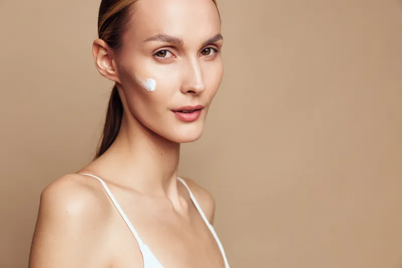 A young woman with clear skin applies moisturizing cream on her cheek, looking confidently at the camera against a beige background. Concept for skincare and beauty routine.