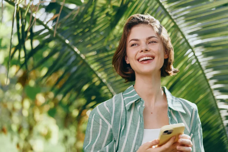Cheerful woman outdoors holding a smartphone, smiling amid lush palm leaves and bright sunlight.