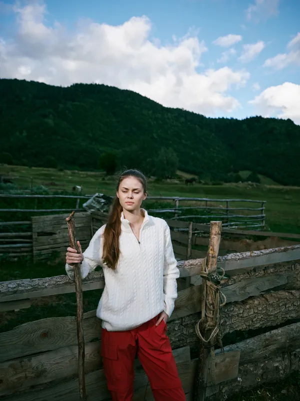 Countryside landscape with a woman holding a wooden staff near a weathered fence, wearing a light sweater and red pants, set against green hills and cloudy sky for rustic outdoor lifestyle