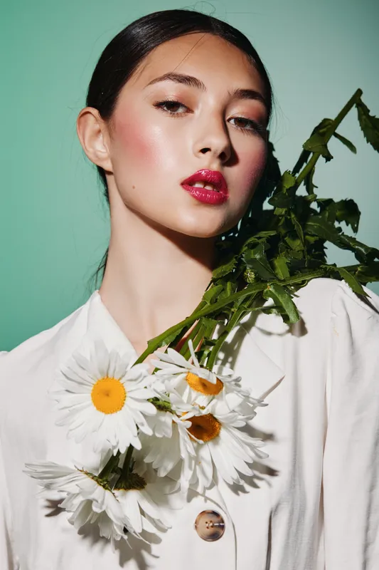 Young woman with glossy red lips and clear glowing skin poses with white daisies on her shoulder against mint background. Concept for beauty creative and fashion innovation.