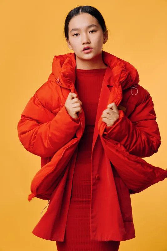 Young woman wearing bright red puffer jacket and matching dress poses against yellow background with serious expression, fashion and winter style concept portrait.