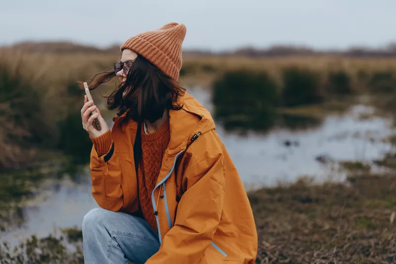 Woman lifestyle outdoors autumn coat beanie landscape river, young adult in orange jacket and jeans, holding cigarette while smoking, relaxed profile walking near stream, natural daylight