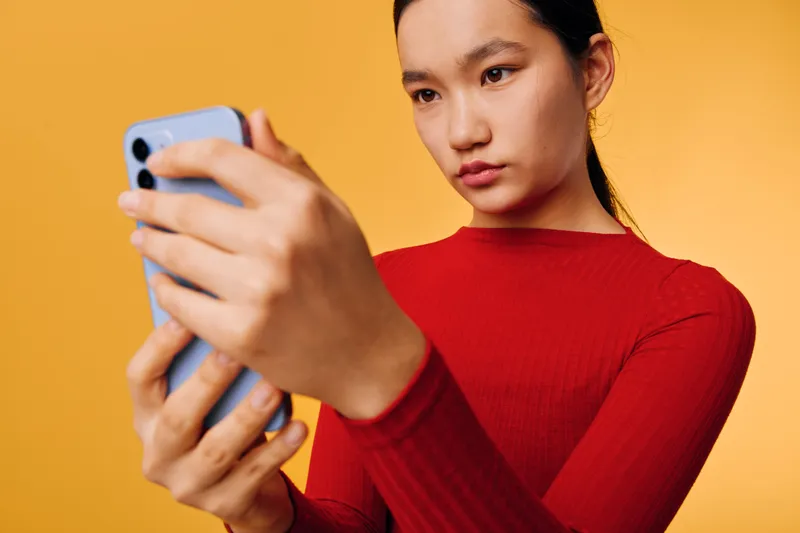 Young woman in red sweater holding smartphone and focusing on screen with serious expression against orange background indoor casual technology communication concept.