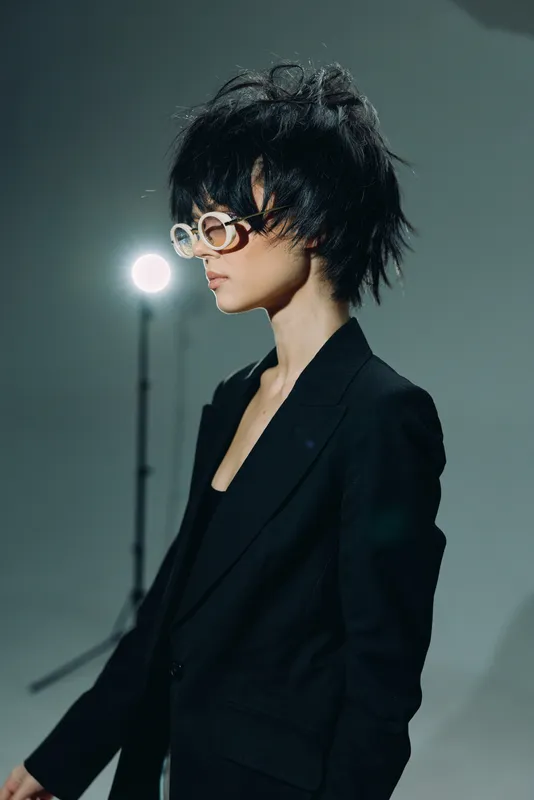 Stylish woman in black suit with short hair and white sunglasses posing against neutral background with studio light