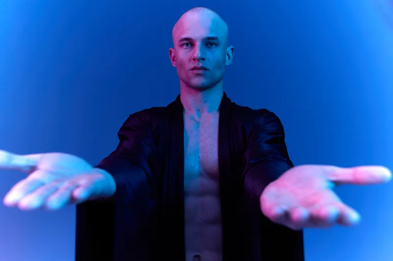 Confident man extending arms in vibrant blue light, showcasing a powerful presence and inviting energy. Unique lighting creates an engaging atmosphere.