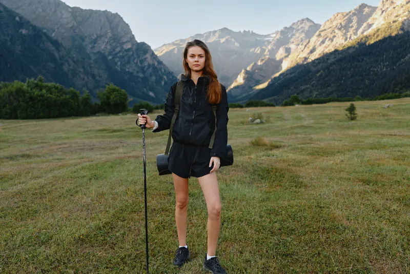 A confident hiker stands in a wide valley, dressed in dark outdoor gear with a trekking pole. She gazes toward rugged peaks, ready for adventure, solitude, and fresh alpine air.