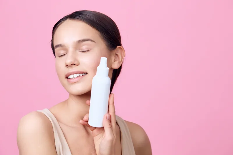 Young woman with closed eyes holding a white skincare bottle, smiling against a pink background, conveying joy and skincare concept