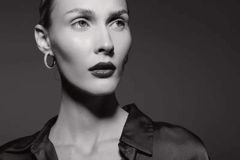 Portrait of a young Caucasian woman with fair skin and short hair wearing hoop earrings and a dark blouse, looking thoughtful in black and white studio lighting.