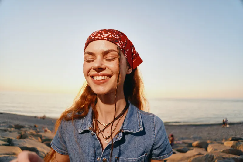 lifestyle woman in modern boho-western denim street style smiling with closed eyes near rocky beach at sunset, film color warm tones vibrant mood