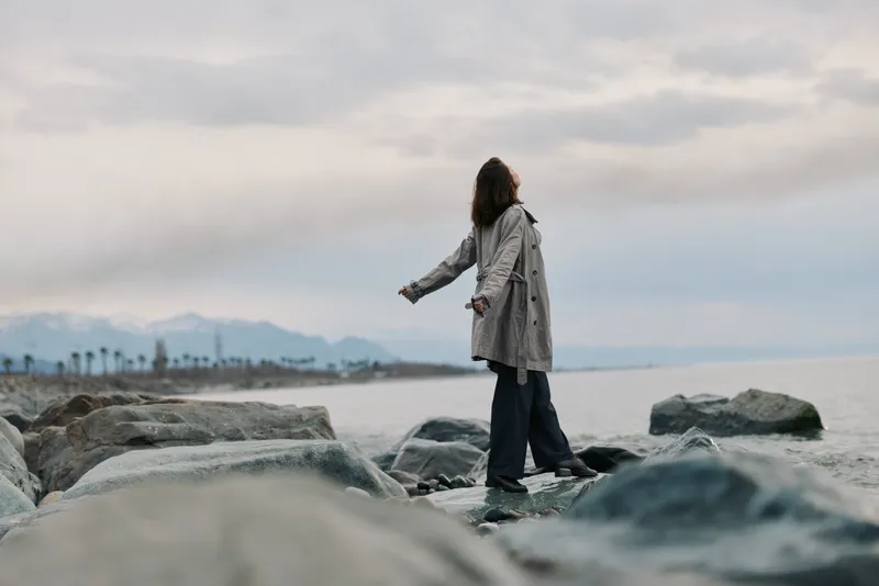 Woman in a long coat stands on rocky shore with arms outstretched, looking at cloudy sky and calm sea. Concept for freedom, nature, and contemplation outdoors.