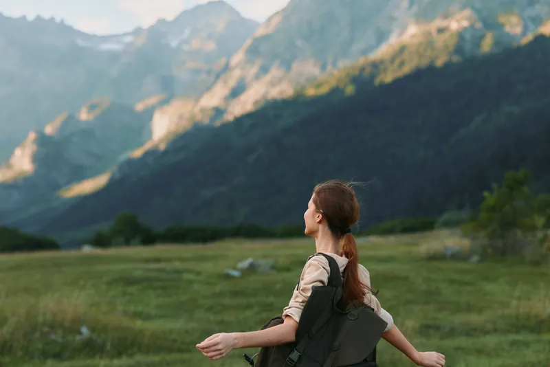 Young woman with a backpack stands in a wide grassy meadow, arms outstretched, gazing toward rugged mountains under clear blue sky, conveying freedom and outdoor adventure.