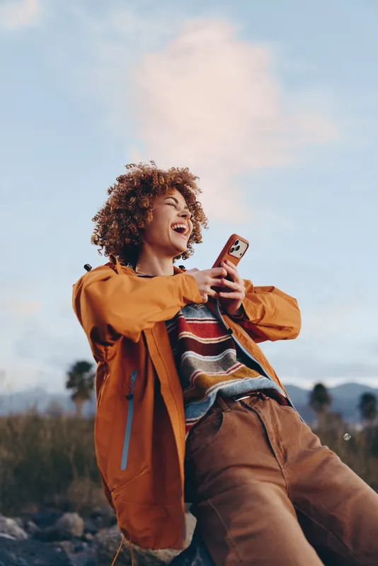 Smiling woman lifestyle in outdoor nature, curly hair and orange jacket, holding smartphone and tapping screen, enjoying fresh air in a scenic field under daylight sky today