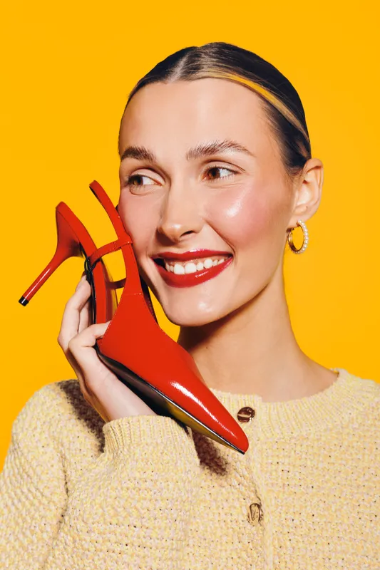 Smiling woman with red lipstick holds red high heel shoe near face on yellow background. Concept of fashion, beauty, style, and happiness with bright studio lighting and cheerful mood.