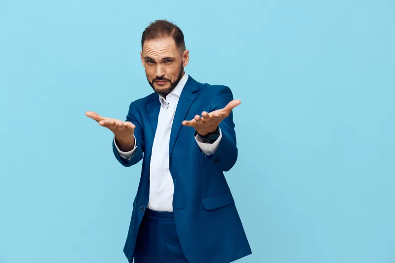 A professional businessman stands before a solid blue background, radiating confidence and focus. His open gesture hints at invitation, curiosity, and readiness to lead.