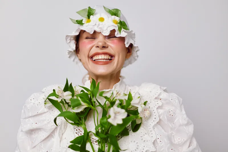 Cheerful woman wearing a flower hat and white dress, holding flowers, exudes happiness and joy, perfect for spring themed concepts, events, and beauty projects