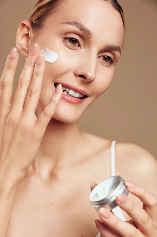 Woman applying face cream, close up on cheek, smiling, concept of skincare and self care, moisturizing beauty routine, holding jar, beige background, soft studio light, wellbeing