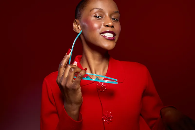 Confident black woman with dark skin wearing red sweater holds blue glasses smiling against red background. Concept of fashion, style, beauty, and positive mood for black female model.