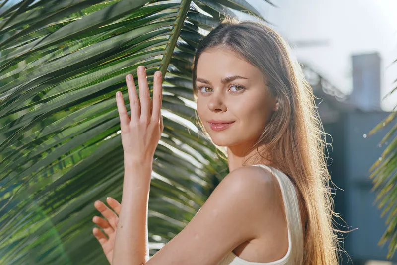 young woman standing by palm leaves outdoors with natural sunlight, wearing a white tank top, looking serene and relaxed in a tropical setting