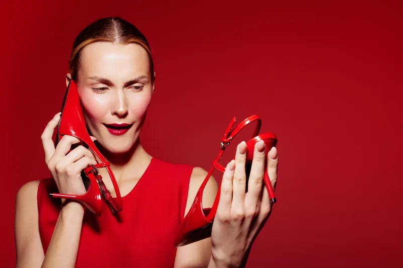 Young woman in red dress holds red high heel shoes, one near ear as if talking, with curious expression on red background. Concept of fashion, style, and playful communication.