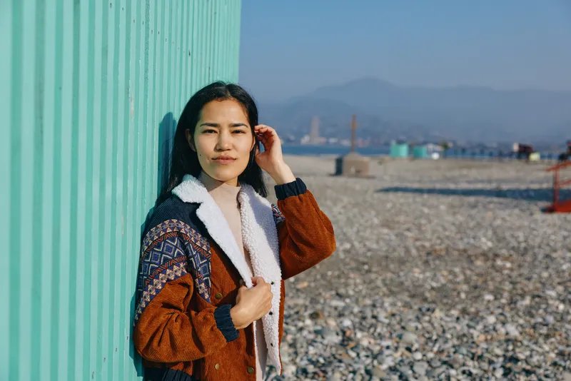Woman beside a turquoise barrier on a pebbled beach, wearing a brown knitted jacket and scarf, gazing at distant mountains, capturing calm coastal scenery and a warm daytime mood