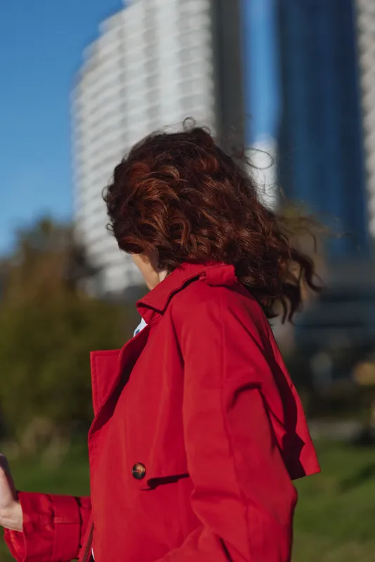 Woman in a red coat outdoors, wind blowing hair, urban skyline in the background. Fashionable street style, dynamic movement, bright colors, warm lighting.