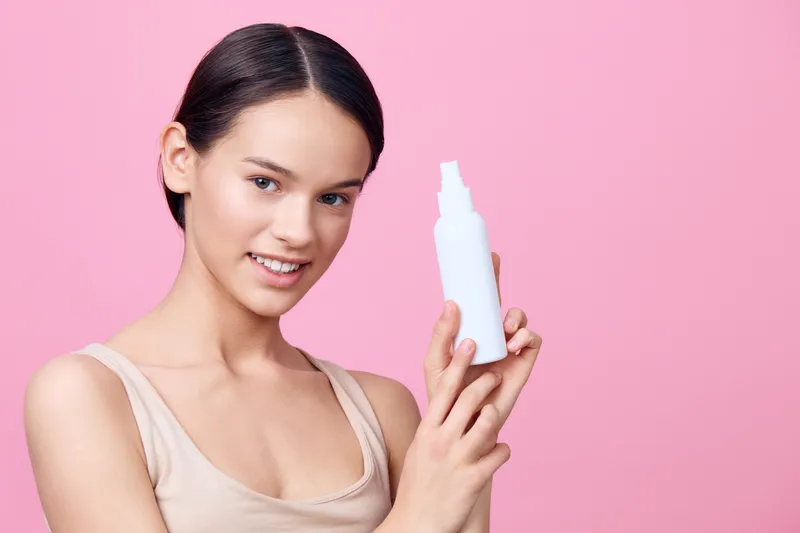 Young woman with a white bottle of skincare product, smiling against a pink background, showcasing a natural beauty and healthy skincare routine