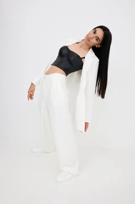 young woman wearing white suit and black top poses leaning backward against plain light background in minimalistic studio setting with casual confident expression