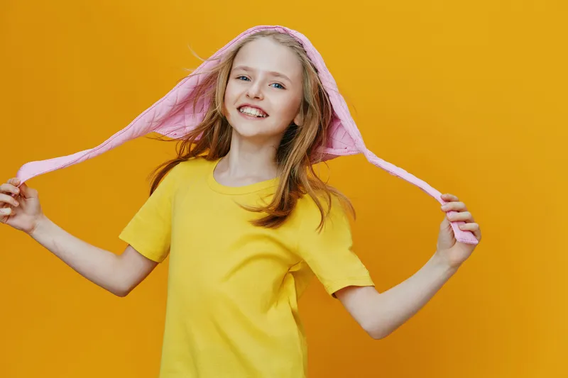 Cheerful young girl in yellow shirt and pink hoodie, smiling against bright yellow background, embodies happiness and playfulness suitable for children s themes