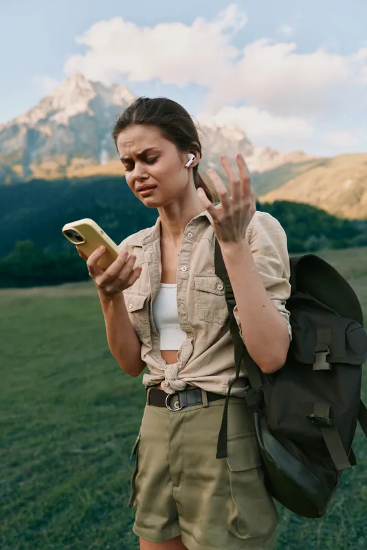Young woman with earbuds looks frustrated while holding a smartphone outdoors in a mountainous area during daytime, wearing casual summer clothes and carrying a backpack.