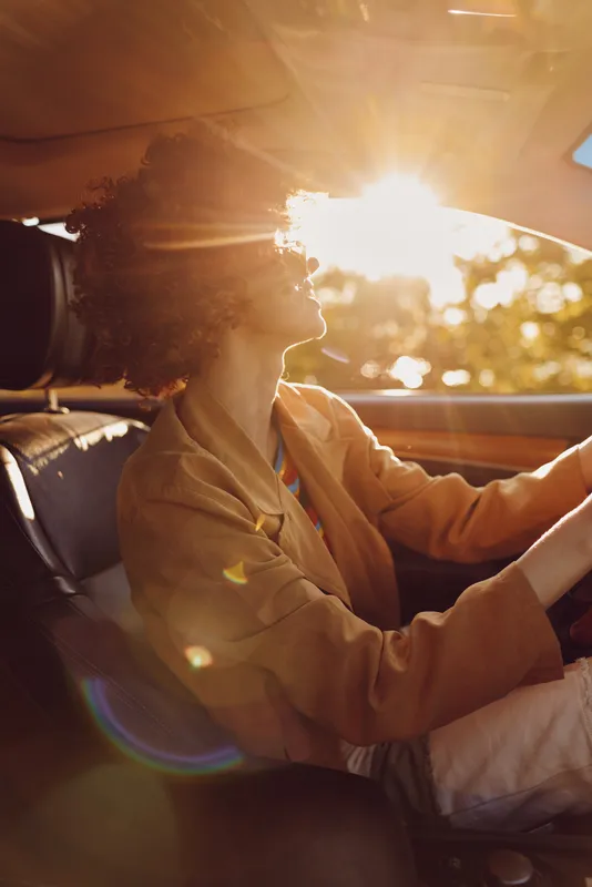 Woman driving a car during golden hour sunset, showcasing retro film color, warm tones, casual lifestyle, inside vehicle, confident expression, candid automotive moment, relaxed atmosphere