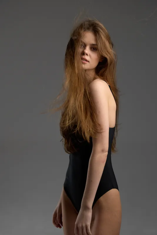 Young woman with long hair wearing a black bodysuit posing against gray background, natural look, studio lighting.
