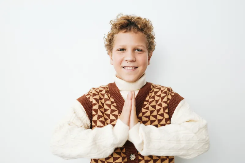 Authentic lifestyle portrait of a young student in a knitted vest, hands pressed in prayer, capturing school spirit, calm authenticity, and a playful, hopeful mood.