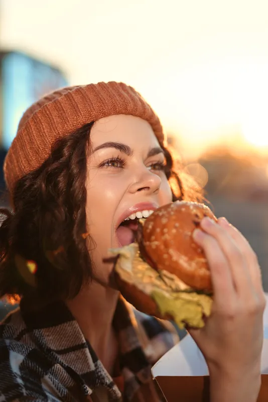 Woman eating burger beanie outdoor candid smile capturing authenticity and emotional storytelling in golden hour glow, mindful living vibe and modern urban lifestyle scene.