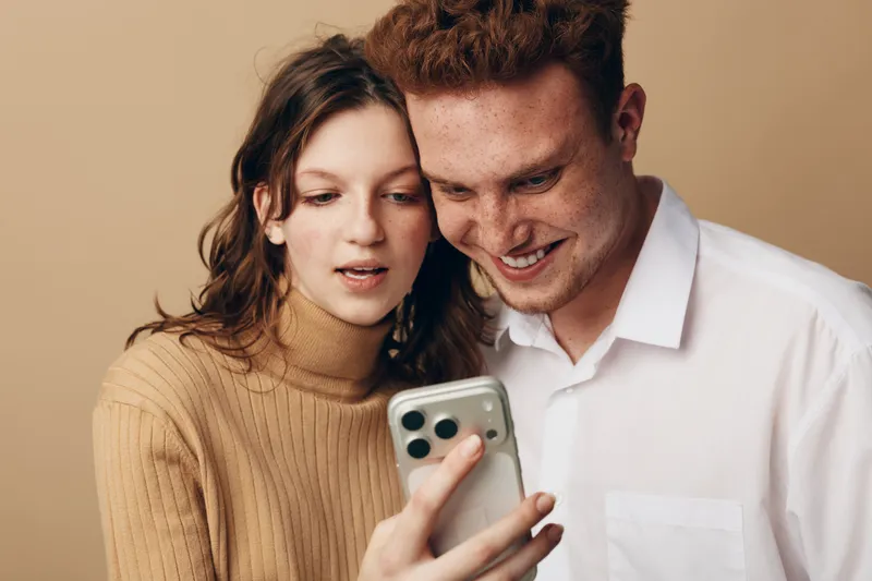 Young couple looking at smartphone screen together, smiling and sharing moment. Casual clothing, indoor setting with neutral background. Concept of technology, communication, and relationship.