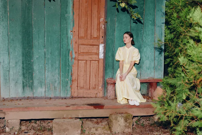 A woman in a pale yellow dress sits quietly on a wooden porch before a weathered door, surrounded by greenery, creating a calm rustic scene with vintage charm.