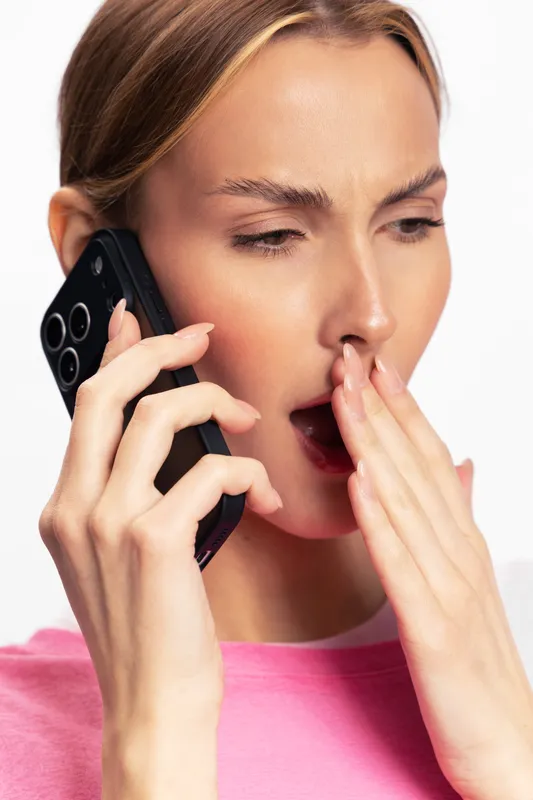 Tired young woman in pink shirt yawns covering mouth while talking on phone. Isolated on white background. Concept of fatigue, communication, and daily routine.
