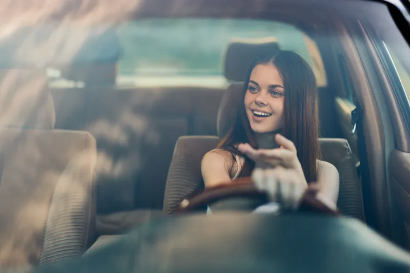 Joyful young woman driving in a car with a bright smile, showcasing freedom and happiness on a sunny day, embodying a carefree spirit and adventure.