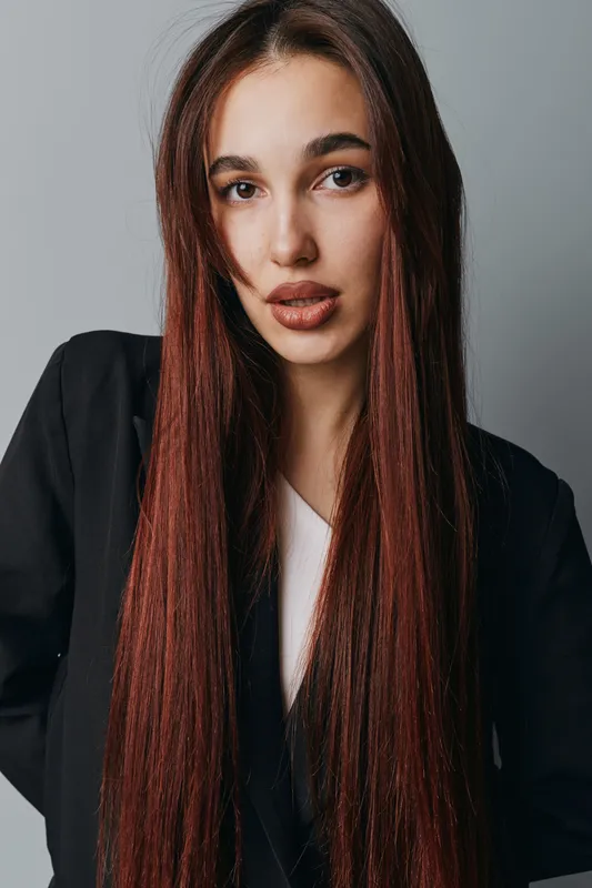 Young woman with long straight hair wearing black blazer and white shirt posing against gray background with confident expression and natural makeup for portrait photography.