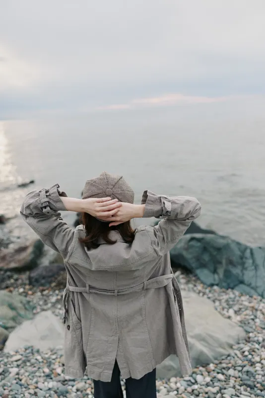 Woman in knitted sweater and hat stands on rocky beach with hands behind head, looking at calm sea under cloudy sky. Concept of relaxation, solitude, autumn, and nature.