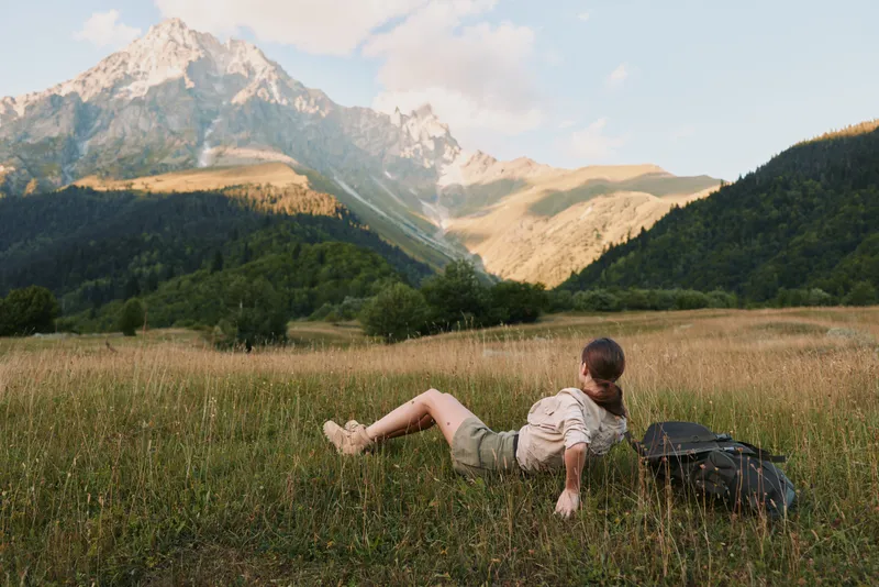 A young woman in casual shorts and a white shirt relaxes on a grassy field with a large backpack nearby while enjoying a mountain view on a clear day.