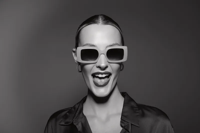 White woman with light skin wearing large sunglasses laughs with open mouth on a grey studio background. Concept of fashion, joy, and confident mood.