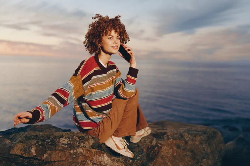 Woman lifestyle on seaside rock at ocean sunset, curly hair, striped sweater and tan overalls, relaxed posing with hand near face, calm moment by shoreline, evening wind, warm colors
