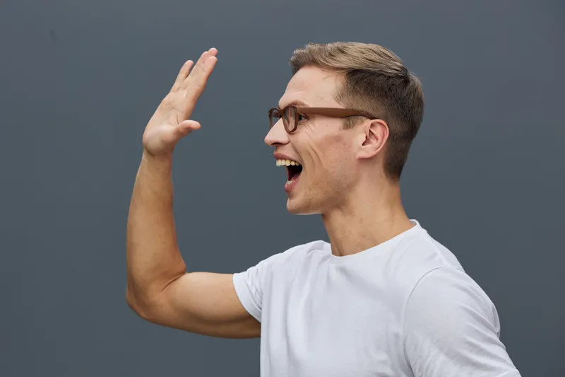 Smiling young man wearing glasses and white t-shirt, expressing happiness and excitement with hand raise, side view, isolated on plain grey background, studio portrait. People lifestyle concept