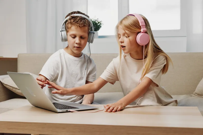 Two children with headphones use a laptop together at home on a beige sofa. They focus on the screen in a bright room. Concept of learning, technology, and collaboration between kids.