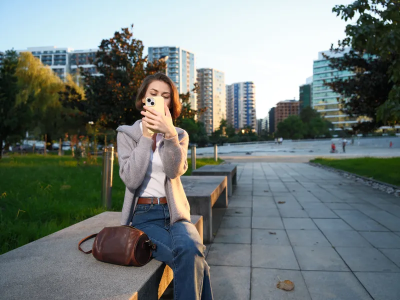 A cheerful woman sits on a concrete bench in a modern urban park, texting on her smartphone. She smiles softly, enjoying a relaxed moment amid city buildings and greenery.