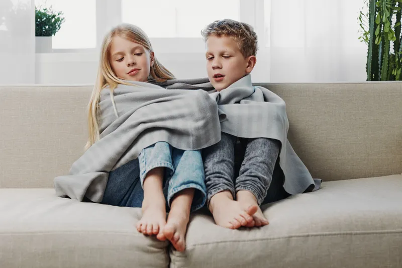 Two children sitting barefoot on a couch wrapped together in a gray blanket, sharing warmth and comfort in a cozy living room with natural light and plants.