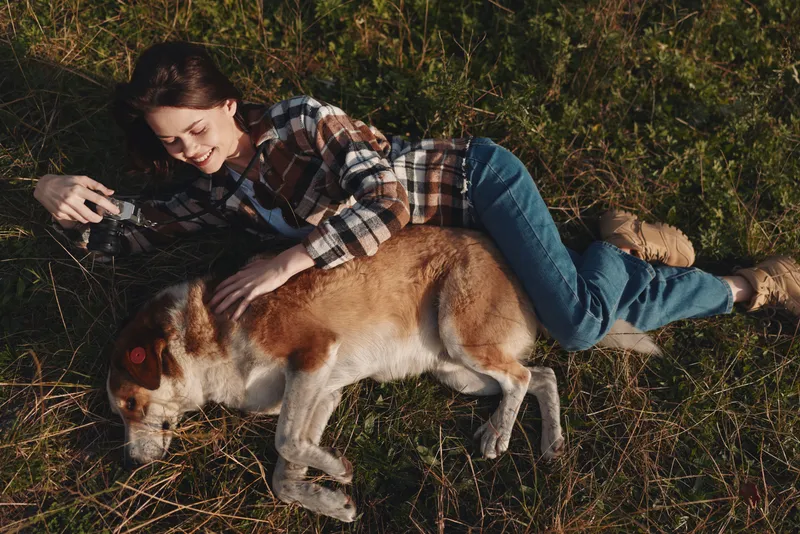 Woman lying on grass beside a calm dog, plaid shirt and jeans, capturing a candid outdoor moment with a handheld camera in natural sunlight