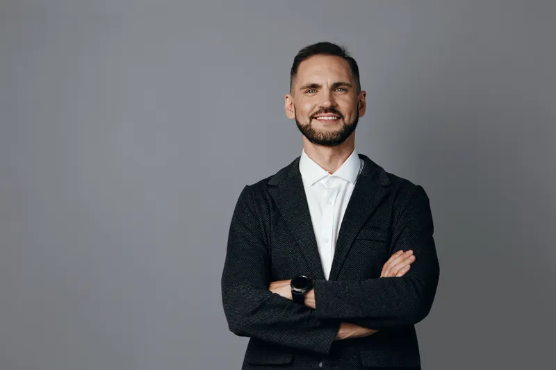 A professional man stands with arms crossed against a solid color background, exuding confidence and warm emotion in a business portrait, with clear presence.