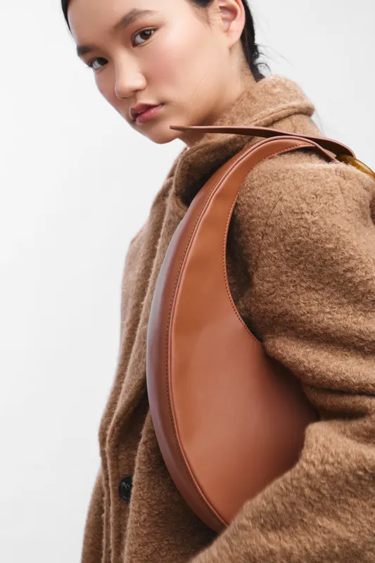 Asian woman wearing a brown coat and leather bag gazes confidently at the camera, presenting a chic fashion moment in a clean studio setting with modern style and poised elegance.